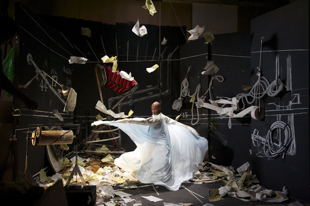 a person in a flowy dress spins amongst a messy room of many objects, their dress is dancing in the air as they move
